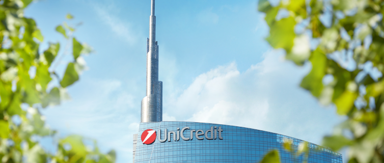 Un moderno grattacielo, la UniCredit Tower, si erge su uno sfondo di cielo azzurro incorniciato da foglie verdi in primo piano. La facciata curva in vetro della torre riflette la luce del sole, enfatizzandone l'elegante design.