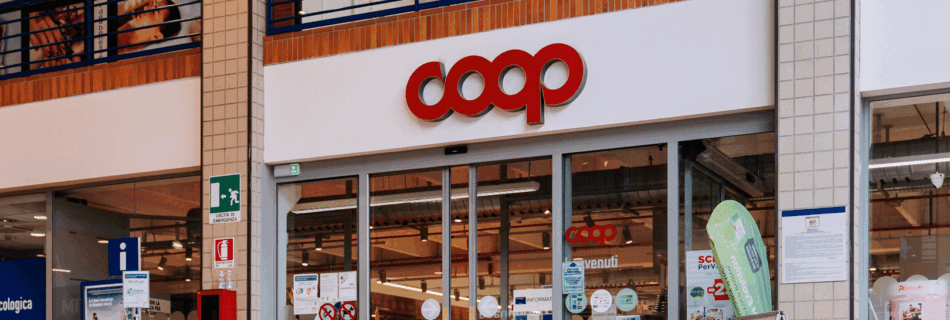 L'ingresso di un supermercato Coop presenta porte in vetro adornate con vari cartelli, tra cui striscioni che avvisano i clienti della truffa TRUFFA COOP che coinvolge falsi buoni sconto. Il nome del negozio è ben visibile sopra l'ingresso ben illuminato, consentendo una chiara visione dell'interno.