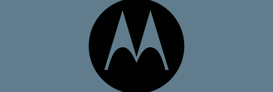 Un cerchio nero contiene una lettera M bianca stilizzata con due punte, che ricordano le ali di pipistrello, centrata su uno sfondo grigio-blu tenue, che ricorda il design elegante della nuova recensione Motorola G84 5G.